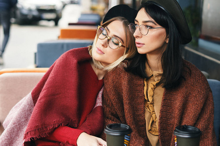 秋天的冬天，两个时髦的女人在户外咖啡馆喝咖啡去摆姿势。 城市背景。 姐妹姐妹姐妹