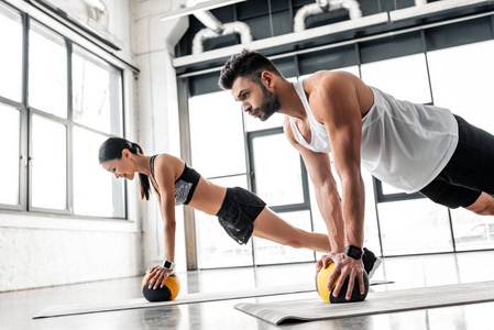 运动青年夫妇在运动服中的侧视，在健身房的瑜伽垫上用药球运动