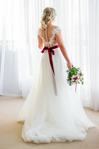 美丽的年轻女孩化妆和发型站在酒店房间里，穿着一件婚纱，手里拿着一束婚礼花束。