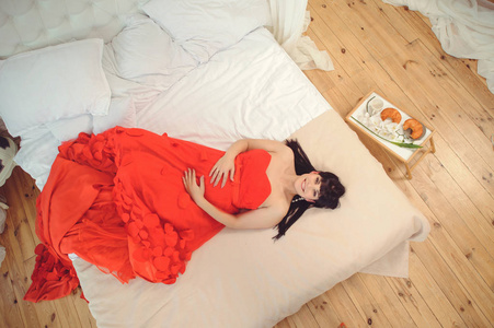新的生活概念。 怀孕孕产和幸福。 特写孕妇穿着时尚优雅的红色连衣裙在床上俯视图像