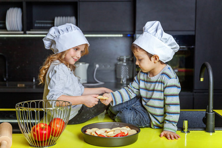 小孩子，女孩和男孩，戴着厨师帽，在厨房准备自制的苹果派。 兄弟姐妹在家做健康的食物，玩得开心。 儿童家庭团队帮助概念