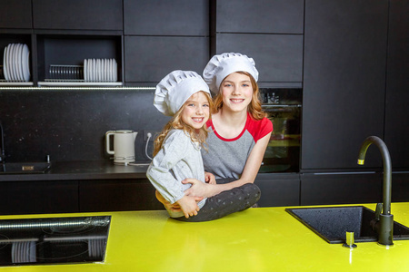 两个女孩带着厨师帽拥抱在厨房里玩得很开心。 姐妹小孩和十几岁的女孩在家做健康的食物。 儿童家庭家庭团队合作帮助概念