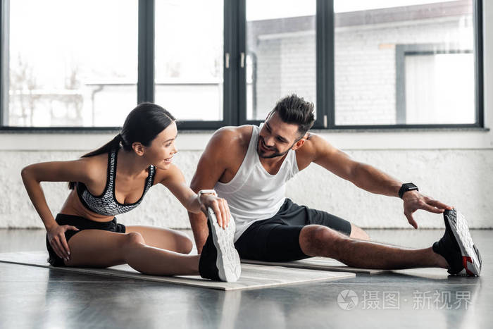 运动的年轻夫妇在健身房训练瑜伽垫时，伸展双腿，互相微笑