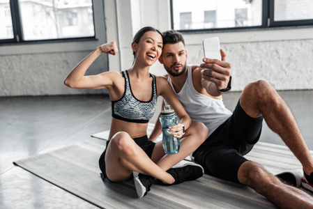快乐的年轻夫妇在运动服上自拍，用智能手机自拍，在健身房锻炼后休息在瑜伽垫上
