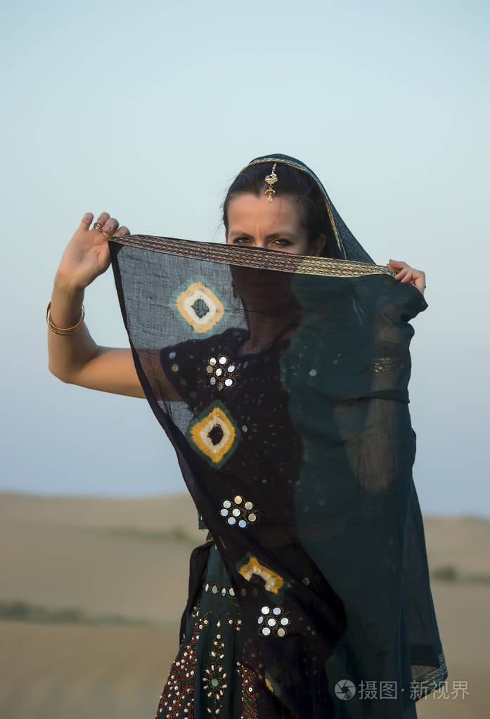 一个穿着印度衣服的女孩把她的脸藏在面纱后面贴在天空和沙丘上