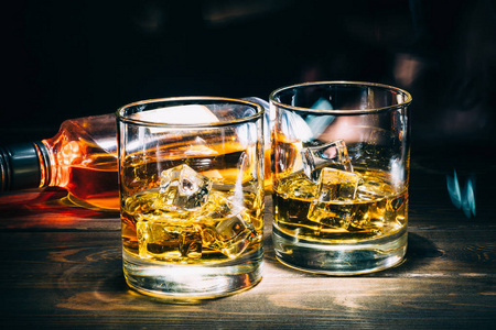 两杯苏格兰威士忌或干邑, 冰块和一瓶酒精酒在黑暗的木制背景