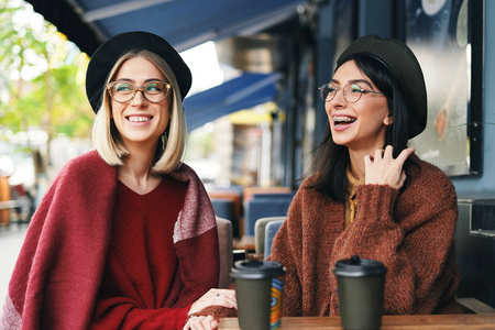 秋天的冬天，两个年轻女人在一家户外咖啡馆喝咖啡去说话。 城市背景。 姐妹姐妹姐妹