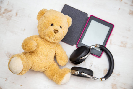 在白色磨损的背景上躺着一本打开的电子书，上面有一个黑色耳机和一个可爱的玩具熊。
