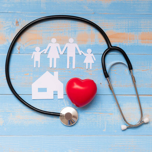 听诊器有红色心形家庭和房子纸上的蓝色粉彩木背景。 医疗及保险概念