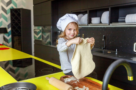 小女孩带着厨师帽，擀面杖，准备面团，在厨房里烤自制的节日苹果派。 孩子在家做健康的食物，玩得开心。 儿童家庭团队帮助概念