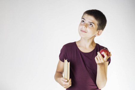 肖像快乐，微笑的男孩拿着书和红苹果，抬头想着什么。快乐的孩子拿着书和苹果。回学校的概念..