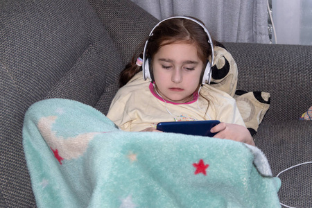 女孩戴着耳机玩电话，躺在沙发上，孩子气的概念玩上瘾，自然的照片