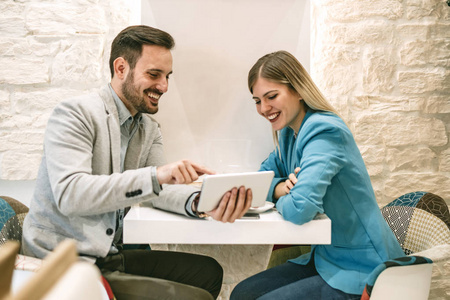 两个美丽的年轻人微笑着坐在咖啡馆里，在数码平板电脑上工作。