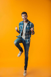 一个穿格子衬衫的快乐年轻人的全长肖像，被隔离在橙色背景上，跳跃庆祝成功