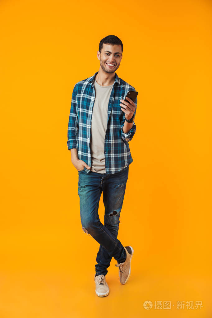 一个穿格子衬衫的快乐年轻人的全长肖像，用手机在橙色背景上隔离