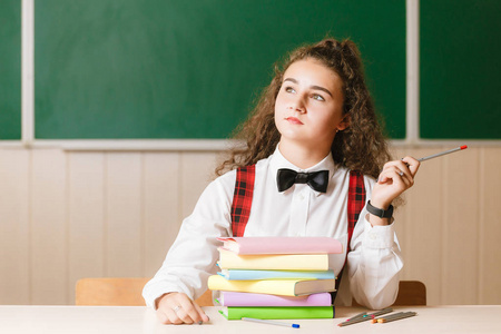 可爱的黑发女学生坐在学校的桌子上，黑板上有书。