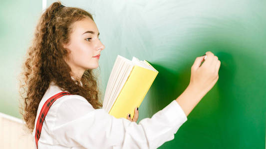 可爱的黑发女学生在黑板上用粉笔写字,上课时手里拿着一本书照片