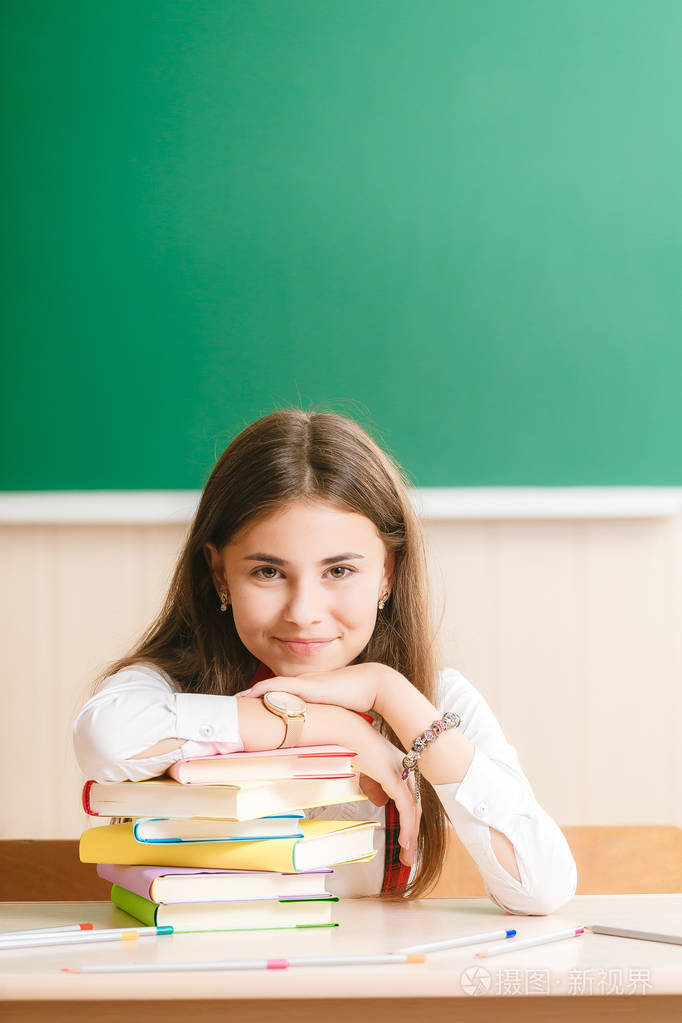 可爱的黑发女学生坐在学校的桌子上,黑板上有书.