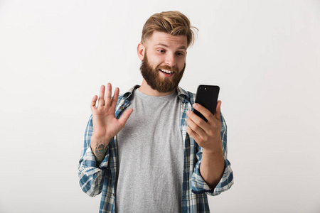 照片上一个英俊的年轻胡须男子站在白色的墙壁背景上，用手机说话挥手。