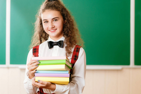 漂亮的女学生站在教室里，在黑板的背景下拿着书。