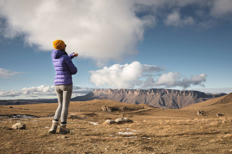 一位女旅行者的背部肖像, 穿着一件带着帽子的夹克, 背包站在一个有岩石的史诗风景的背景上