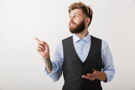 照片上一个英俊的年轻胡须男子站在白色墙壁背景上，使用手机指向。