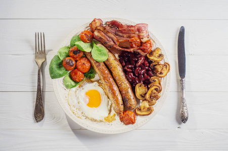 美味的丰盛的英式早餐，包括煎鸡蛋培根香肠西红柿豆类和蘑菇。 传统英式早餐在白色木制背景上，俯视平面。