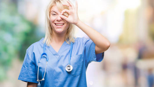 年轻漂亮的金发医生女人穿着医疗制服，在孤立的背景下做着OK的手势，用手微笑的眼睛透过手指看着幸福的脸。