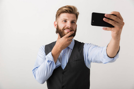 一个英俊的年轻胡须男站在白色墙壁背景上，用手机自拍。