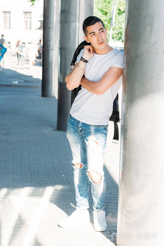 年轻的帅哥穿着一件白色的t恤,黑色的皮夹克和蓝色的破牛仔裤,在街上