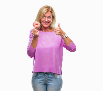 中年金发女人吃粉红面圈在孤立的背景上快乐的大笑着做，好的，用手指签拇指，好的标志