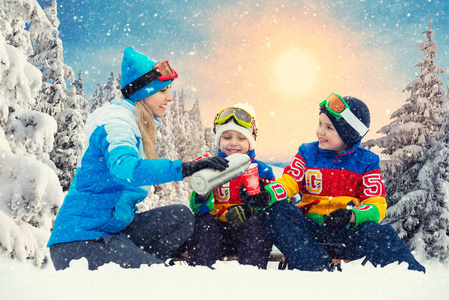 母亲和两个儿子在雪橇后在雪林里喝热茶。 一起喝热茶。圣诞节假期的家庭冬季乐趣。