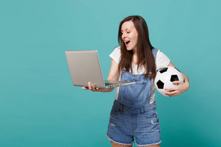 开朗的年轻女子足球迷拿着足球，使用笔记本电脑电脑，隔离在蓝色绿松石墙背景上。 人们情感运动家庭休闲生活方式的概念。 模拟复制空间