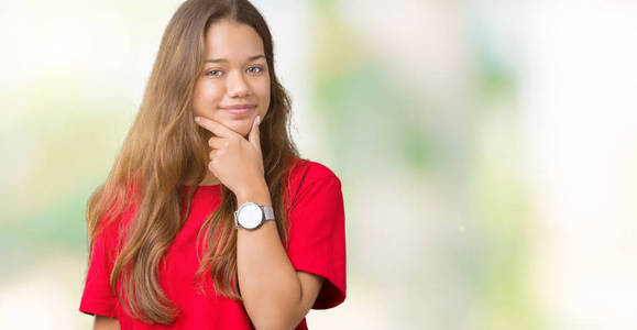 年轻漂亮的黑发女人，穿着红色T恤，在孤立的背景下，自信地看着相机，微笑着交叉的手臂和举起的手在下巴上。 积极思考。