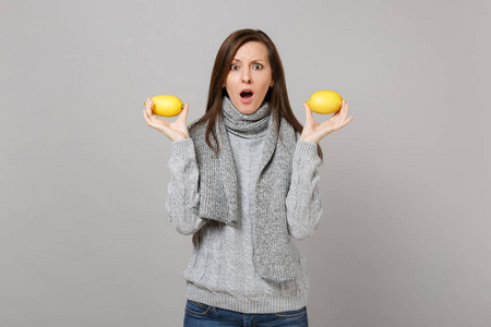 震惊的年轻女子穿着灰色毛衣围巾,嘴巴张开,把柠檬隔离在灰色背景上.