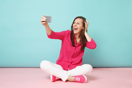 全长女性穿着玫瑰衬衫，白色裤子，坐在地板上，在手机上自拍，隔离在明亮的粉红色蓝色粉彩墙背景工作室。 时尚生活方式的概念。 模拟复