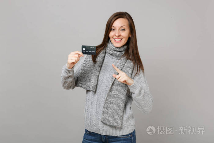 年轻女子戴着灰色毛衣围巾，食指指向信用卡上的灰色墙壁背景。 健康时尚生活方式人真诚情感冷季观念。 模拟复制空间