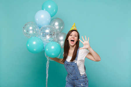 有趣的年轻女子戴着生日帽，张开嘴，露出手掌，庆祝手持五颜六色的气球，孤立在蓝色绿松石背景上。 3.生日派对4.情感观念