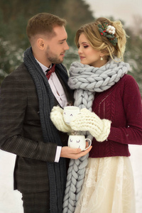一对夫妇，一个男人和一个女人站在冬天的森林里，手里拿着杯子，穿着毛衣和西装，戴着一条大针织围巾。