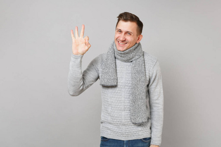 快乐的年轻人穿着灰色毛衣围巾，在灰色背景的工作室肖像上展示了OK手势。 健康时尚生活方式人真诚情感冷季观念。 模拟复制空间