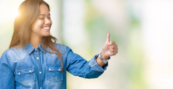 年轻漂亮的黑发女人，穿着蓝色牛仔衬衫，在孤立的背景下，看上去骄傲地微笑着，竖起大拇指，向旁边做手势。