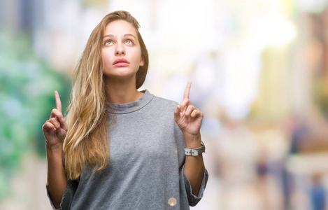 年轻漂亮的金发女人在孤立的背景上惊讶和惊讶地抬头，用手指和举起的手臂指着。