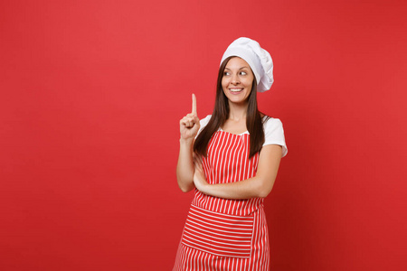 家庭主妇女厨师或面包师穿条纹围裙，白色T恤，烤厨师帽子隔离在红墙背景上。 美丽的管家女人指着食指。 模拟复制空间概念