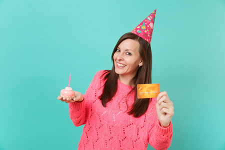 微笑，美丽的年轻女子，穿着针织粉色毛衣，生日帽，手拿蛋糕，蜡烛信用卡隔离在蓝色绿松石墙背景上。 人们的生活方式概念。 模拟复制空