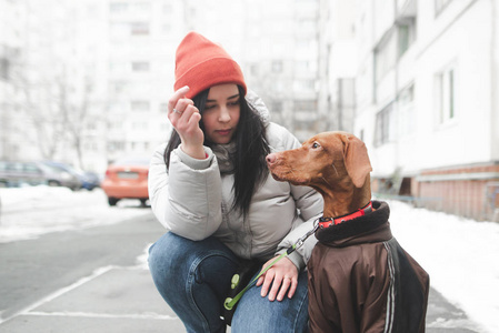 美丽的微笑女孩穿着温暖的冬天衣服，在街道和建筑物的背景上养了一只穿着衣服的狗。 冬天和一只狗在院子里散步。 专注于一个有一只漂亮