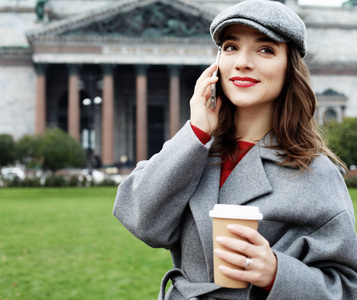 一个漂亮的微笑的女人的肖像使用手机, 而在城市街道上拿着咖啡杯