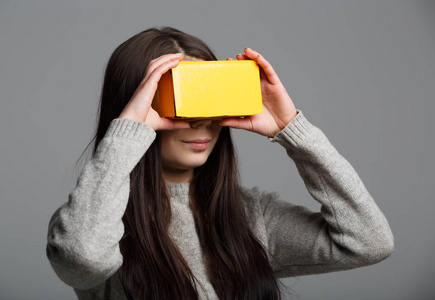 年轻女孩在虚拟现实耳机设备上玩手机游戏应用程序。用于移动游戏的黄卡板盒。游戏玩家可携带的VR头套技术。玩移动游戏的增强现实小工具