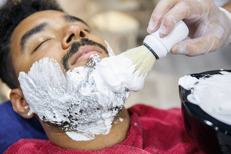 理发师在理发店沙龙用拭子刷在年轻的黑人脸上涂抹白色剃须膏。 特写中的男性美容治疗过程