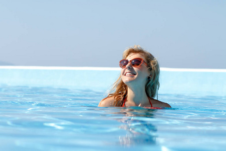 迷人的年轻金发女人在明亮的夏天阳光下在干净的蓝色酒店游泳池里游泳。 漂亮的模特儿