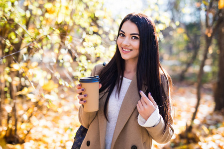 秋天的女人在秋天的城市公园喝咖啡。 享受一次性咖啡杯热饮的女孩。